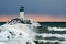 Lighthouse at lake ontario in winter with pink horizon, blue cloudy sky and waves crashing on the rocks