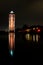 Lighthouse tower next to an enormous lake at night with street lanterns and reflection in the water