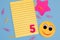 A lined yellow Notepad with the number five, pink star sticker, scattered paper clips and a smiling yellow smiley face