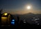 Lit-up camps on a campground with a rising full moon on a hill top in the background. Trekking and camping in the Himalayas.