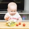 Litlle infant caucasian boy is trying to steal one unhealthy delicious hotdog with green salad from the table