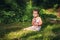 Little 3 years old short haired girl in gray dress sits on green grass in garden with small pink elephant toy. Sad child in summer