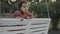 Little 5 yo girl having fun on a white swing in a quiet, tree-lined park background. Preschooler relaxing on a bench