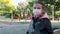 Little adorable caucasian girl in face mask stands outdoor look at camera, on background closed playground park area, due corona