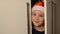 Little adorable girl child in a Christmas red santa claus hat peeks out from behind the cabinet door