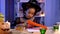 Little African American girl in festive costume and witch hat cuts pumpkin out of orange paper. Child sits at table in