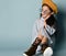 Little asian child in sunglasses, shirt dress, brown beret, boots. Smiling, posing sitting on blue background. Close up