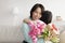 Little Asian girl hugging granny, presenting her with flowers and gift for Women's Day, congratulating with birthday