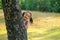 Little Asian girl play peekaboo by hiding back of tree and smile, she play in green garden with morning light