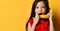 Little asian kid in red blouse. She pretending to be talking on banana like by phone, posing on orange background. Close up