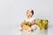 Little babe in dress and headband, barefoot. She posing with two golden gift boxes, sitting on floor  on white. Close up