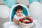 Little baby boy sitting in high chair in white kitchen and tasting first year cake with fruits on background with balloons