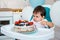 Little baby boy sitting in high chair in white kitchen and tasting first year cake with fruits on background with balloons