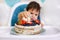 Little baby boy sitting in high chair in white kitchen and tasting first year cake with fruits on background with balloons