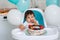 Little baby boy sitting in high chair in white kitchen and tasting first year cake with fruits on background with balloons