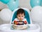 Little baby boy sitting in high chair in white kitchen and tasting first year cake with fruits on background with balloons