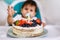 Little baby boy sitting in high chair in white kitchen and tasting first year cake with fruits on background with balloons