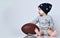 Little baby boy toddler in grey casual jumpsuit, black cap with stars and barefoot sitting on floor with rugby ball