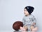 Little baby boy toddler in grey casual jumpsuit, black cap with stars and barefoot sitting on floor with rugby ball