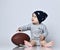 Little baby boy toddler in grey casual jumpsuit, black cap with stars and barefoot sitting on floor with rugby ball