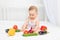 Little baby girl 6 months old sitting on the bed in the nursery with vegetables, baby`s first feeding, baby food concept, place
