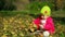 Little baby girl sits motionless on grass and looks at the falling leaves in park on bright and sunny early autumn day.Little baby