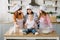 Little baby girl is sitting on the wooden table at kitchen while her mother, aunt and grandmother read the book with