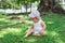 Little baby girl in white clothing and panama is playing with flowers on grass in park at summer. Child explores world around
