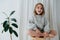 Little barefoot girl in a sweater sitting cross-legged on a stool at home