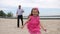 A little, beautiful girl running with her grandfather on the beach. The girl runs on the foreground, the grandfather in