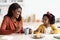 Little Black Girl And Her Mom Eating Snacks And Chatting In Kitchen