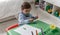 A little blond boy with a pacifier and dressed in blue, plays with some crayons on a small desk, while he is in his bedroom