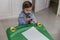 A little blond boy with a pacifier and dressed in blue, plays with some crayons on a small desk, while he is in his bedroom