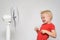 Little blond boy in a red T-shirt makes a face near the fan. Summer concept