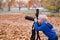 Little blond boy shoots with a large SLR camera on a tripod. Photo session in the autumn park