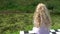 Little blond girl with curly hair sitting on checkered bench near pond in park