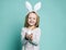 Little blond girl in gray cosy home dress and decorative fur ears standing, holding pastel blue Easter egg in hands