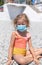 Little blond girl in a swimsuit and a protective medical mask on a pebble beach.