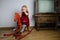 Little blonde girl 2 years old in a red velvet dress sits on a wooden old rocking horse in the room by the old TV, blank screen,