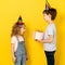 Little blonde girl accepts gift in pink box for her birthday from her brother, on a yellow background, children in caps