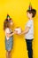 Little blonde girl accepts gift in pink box for her birthday from her brother, on a yellow background, children in caps