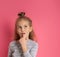 Little blonde girl with bun hairstyle, in gray striped blouse. Put forefinger in mouth, thoughtful, posing on pink