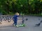 Little blonde girl carries a green scooter past a flock of pigeons. City park in the summer afternoon