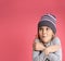 Little blonde girl in gray striped blouse and hat. She hugging herself, looking frozen, posing against pink studio