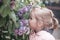 Little blonde girl in the park smells lilac flowers on a tree. Allergy to pollen. The concept of childhood and spring holiday