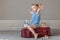 little blonde girl sitting on burgundy suitcase with transparent full money dollars, arms outstretched, playing airplane