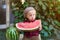 A little boy with blond hair n the summer on a sunny day sitting at a gazebo with a green grapes and eating a watermelon