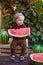 A little boy with blond hair n the summer on a sunny day sitting at a gazebo with a green grapes and eating a watermelon