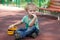 Little boy with blue eyes sits on the cover of children playground with a toy - yellow excavator. Blonde hair, thoughtful look, al