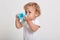 Little boy with blue plastic baby cup drinking water, thirsty child in t shirt posing isolated over white background, looking away